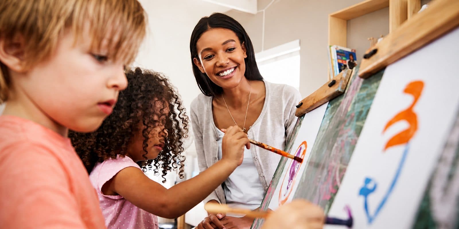 A teacher supporting two young children painting at an easel